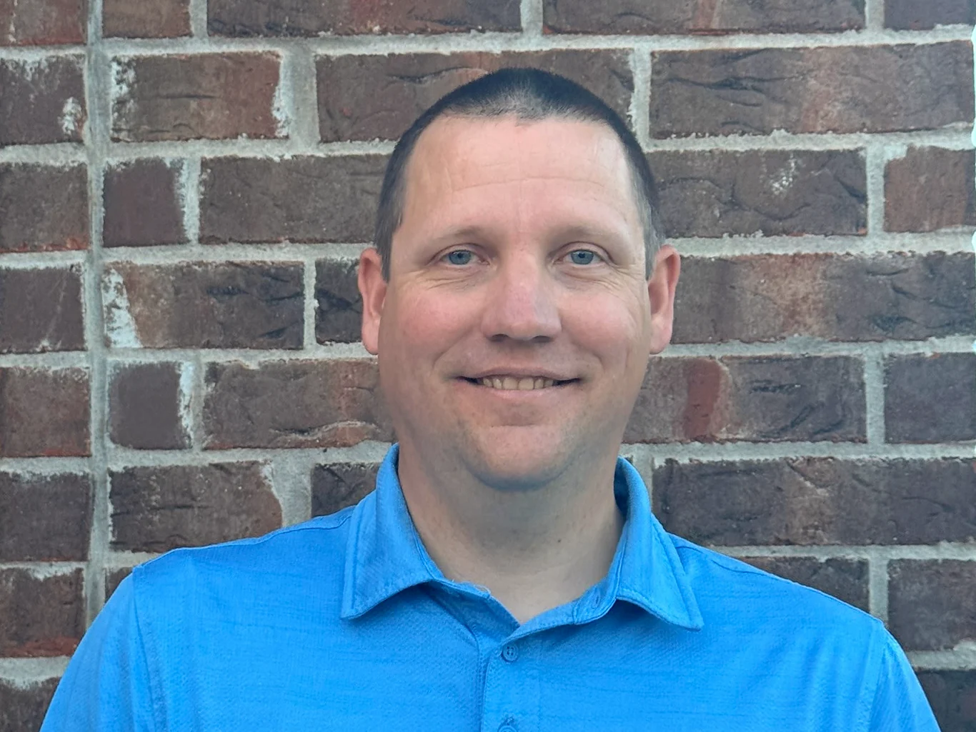Justin smiling in a blue polo shirt against a brick wall